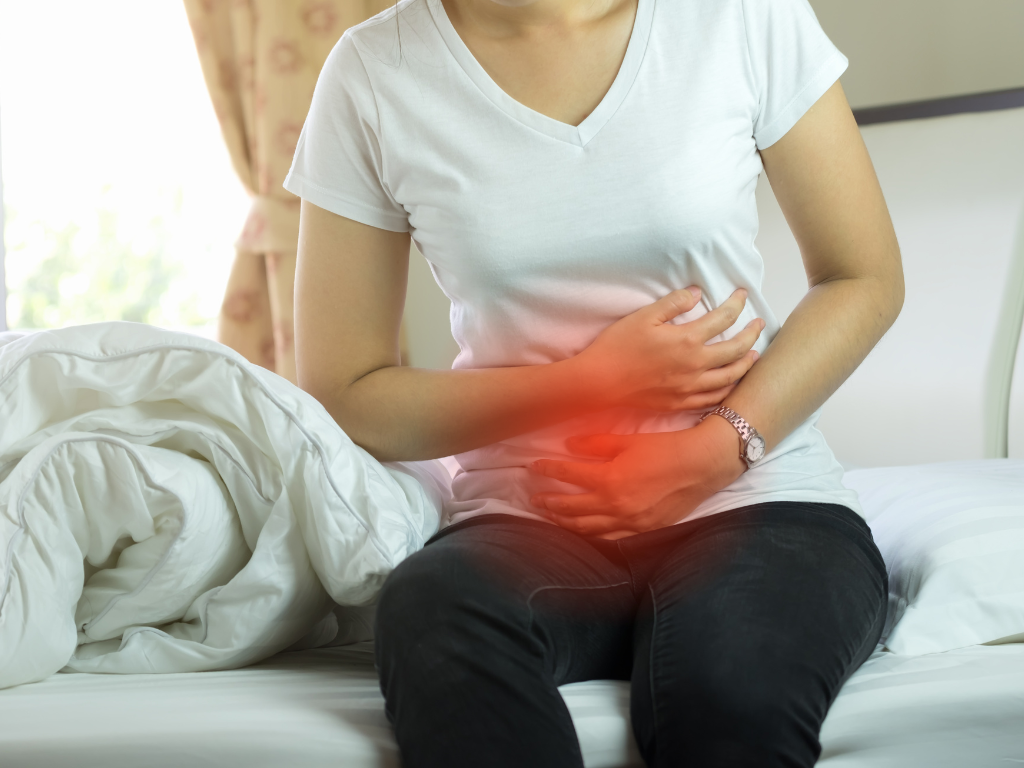 Mujer con dolor abdominal asociado a inflamación silenciosa, sentada en la cama y sujetándose el estómago.